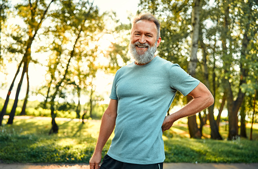 Sport during leisure. Portrait of caucasian pensioner in light blue t-shirt smiling and looking at camera while standing at summer park. Healthy fit man with grey hair resting after outdoors workout.