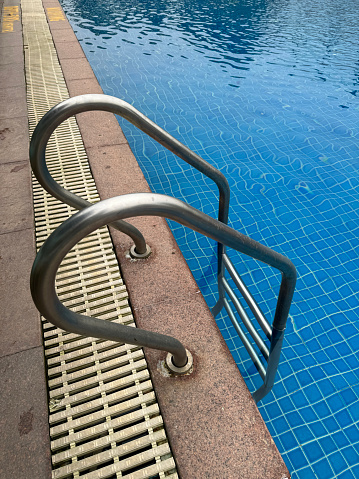 Stock photo showing close-up view of swimming pool with turquoise blue mosaic tiles lining the length of the pool. Metal hand-railed steps enable people to access the water easily and safely.