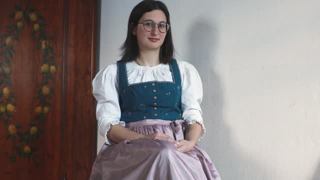 A model wearing a traditional antique Austrian dirndl dress with a pink apron and embroidery on the bodice sits on a chair near a painted wardrobe, Salzburg, Austria