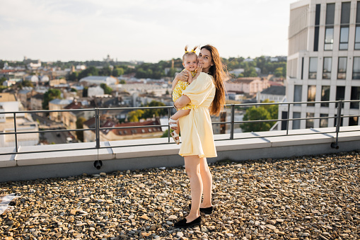 Dark haired woman wearing summer dress standing and holding happy baby in arms while hugging. Caucasian young mother educating and showing kid views from roof of high-rise building.