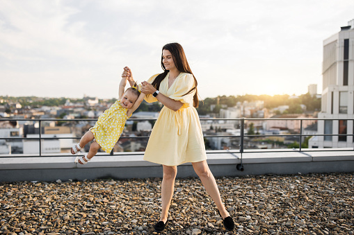 Beautiful smiling brunette holding little daughter in arms and spinning on roof of high-rise building. Cheerful adult lady wearing summer yellow dress enjoying happy moments with lovely child outdoors