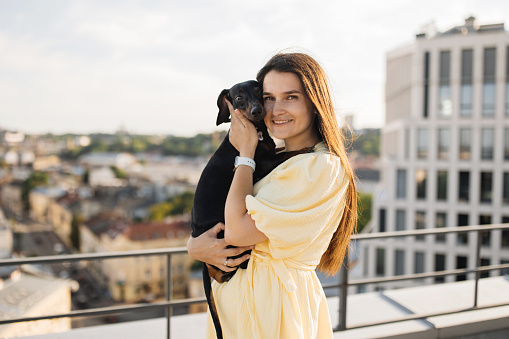 Portrait of cheerful woman with long dark hair standing on rooftop and embracing black dog in arms. Caucasian young female walking with lovely pet during warm summer day.