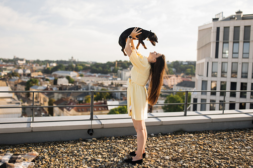 Side view of tall beautiful female standing on roof of high-rise building and holding up black dachshund in hands. Caucasian brunette relaxing and enjoying moment with pet in fresh air.