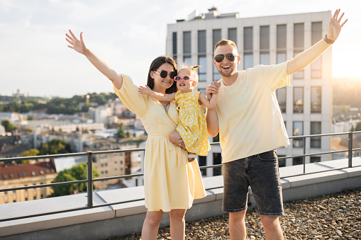 Attractive young man and woman raising hands while holding little daughter. Happy caucasian parents with lovely kid standing on rooftop with white high building on background.