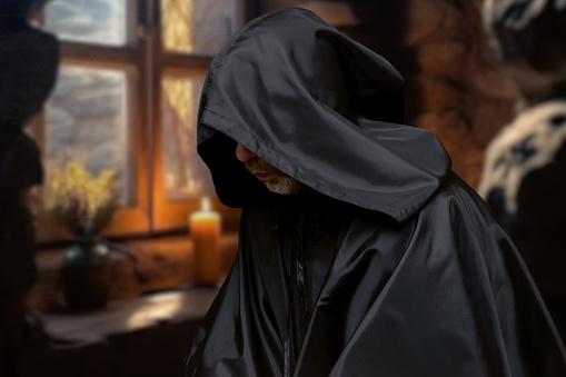 Elderly monk in black cassock praying in a cell in front of a window with a candle