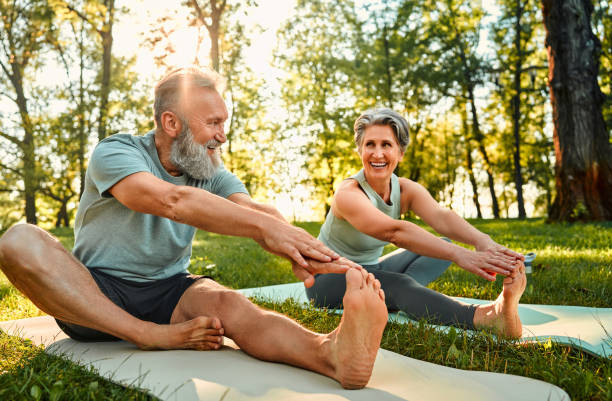 flexible übungen für den körper. sportlicher mann und frau mit grauen haaren strecken sich auf yogamatten mit den händen an einem bein während des outdoor-trainings. glückliches ehepaar mit nackten füßen wärmt sich gemeinsam im park auf. - senior adult sport yoga exercising stock-fotos und bilder