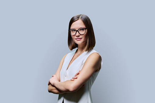 Young confident trendy smiling woman in eyeglasses with crossed arms looking at camera on gray studio background. Beauty, business, work entrepreneurship business expert owner agent concept
