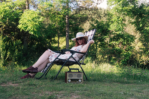Young woman on the chair in nature listening to the radio and relaxing, wearing dress and sunglasses and a hat on a summer day.