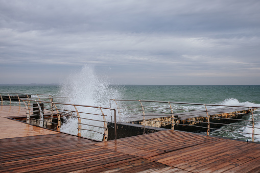 a sea wave breaks on a pier with a wooden deck in cloudy weather