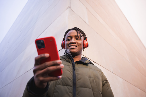A man is listening to music with his smartphone and headphones, leaning on the wall. Young adult disconnecting from the world and connecting with their own thoughts and feelings by immersing themselves in music in an urban scene.