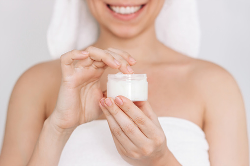 Cropped shot of a young smiling woman in a white towel after shower taking moisturizing cream with the finger holding cosmetic jar in her hand on a light background. Skin care. Cosmetology and beauty