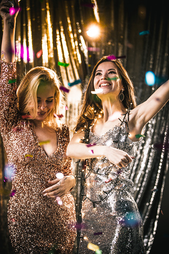 Two girls at the New Year's Eve party blow up firecrackers, a sequin dress. Having fun at the holiday. A young smiling blonde and brunette woman in the studio with confetti. Celebration concept.