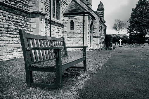 Shallow focus of a wooden tribute bench seen outside a typical English church. The path leads to a highlight stone cross in the distance.
