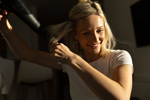 A young blond woman is drying her hair at her home. She is smiling, looking happy.