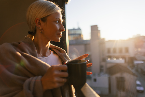 A young woman drinking coffee on the balcony. It's early morning, and it's cold. She is wrapped in a blanket, looks relaxed.