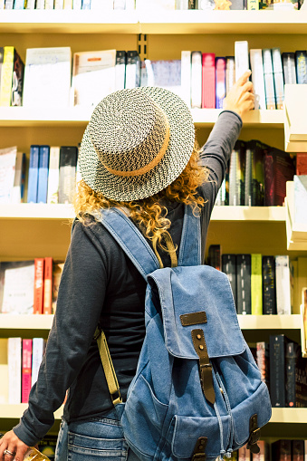 Back view of woman with hat and backpack choosing a book to buy inside a library or newspaper store. People prepare to travel. Leisure and shopping female people activity. Traveler lifestyle