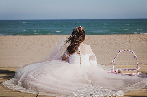 Girl on her back sitting on the beach in her communion dress and looking at the sea.