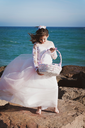 Happy little girl with a basket and her Communion dress, standing on the rocks on the beach.