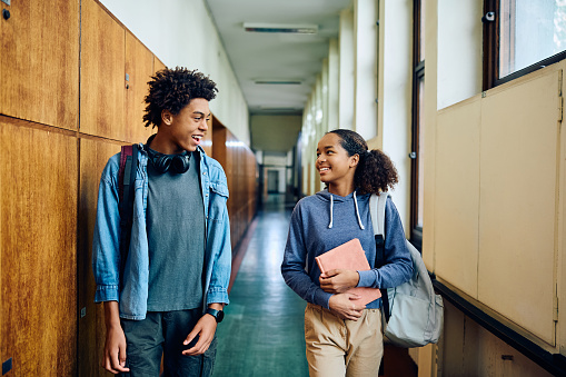Happy African American teenage girl and her classmate communicating while walking through hallway at high school. Copy space.