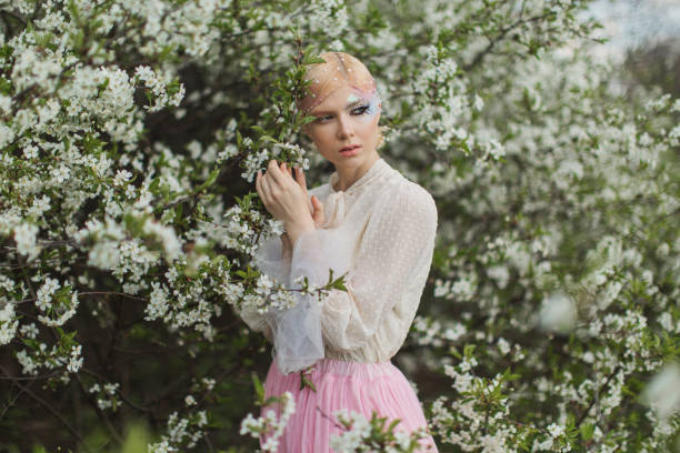 istock Blonde woman with beauty makeup with feather on face. White cherry blossoms. Spring tree background. 1912541634
