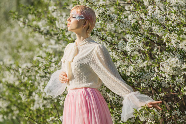 istock Blonde woman with beauty makeup with feather on face. White cherry blossoms. Spring tree background. 1912567995