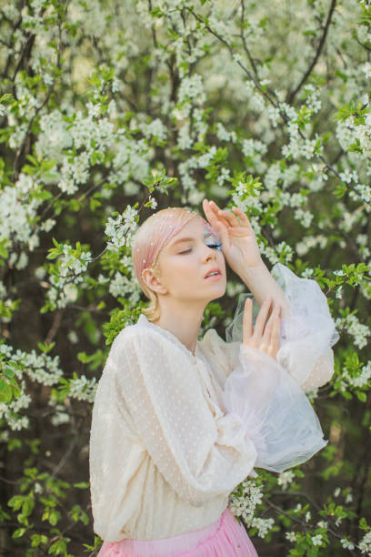 istock Blonde woman with beauty makeup with feather on face. White cherry blossoms. Spring tree background. 1912578088