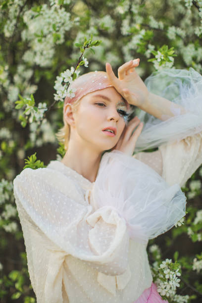 istock Blonde woman with beauty makeup with feather on face. White cherry blossoms. Spring tree background. 1912591062
