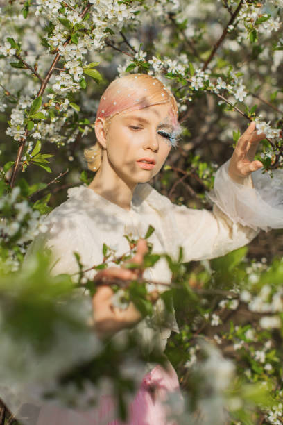 istock Blonde woman with beauty makeup with feather on face. White cherry blossoms. Spring tree background. 1912607011