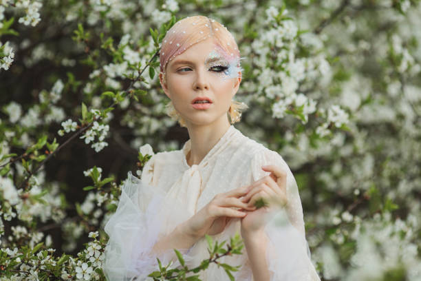 istock Blonde woman with beauty makeup with feather on face. White cherry blossoms. Spring tree background. 1912618016