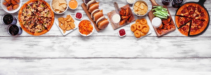 Junk food top border. Pizza, hamburgers, chicken wings and salty snacks. Above view over a white wood banner background.