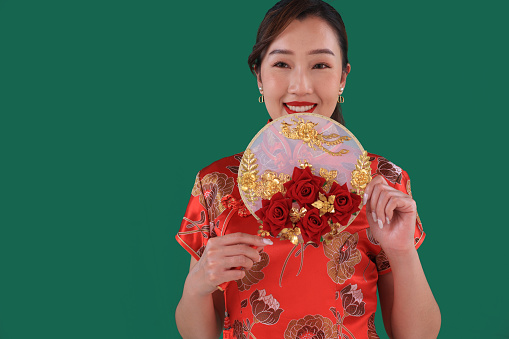 A cheerful Asia woman with a traditional cheongsam qipao dress holding a fan with a smile against a green background