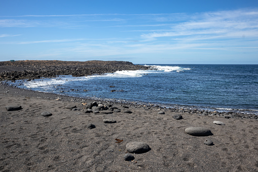 Beach with a dark sand, created from lava. Big pebbles. Calm water of the Atlantic ocean. Blue sky with light white clouds. Lanzarote, Canary Islands, Spain.