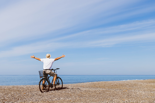 Carefree man standing on the seashore with arms wide open. Happy tourist with bicycle enjoying the sea view. Travel concept. Copy space.