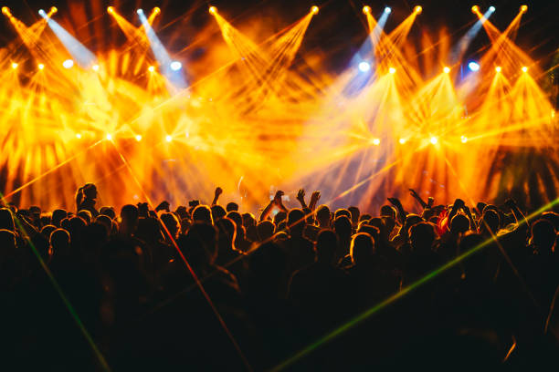 Silhouettes of people dancing and rising hands at open air summer festival. Crowd is enjoying live performance at open air summer festival at night. Nightlife. Silhouettes of people are standing in the dark and listening to electro music at concert. There are orange lights. crowd of people stock pictures, royalty-free photos & images