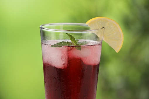 Refreshing hibiscus tea with ice cubes, mint and lemon in glass against blurred green background, closeup