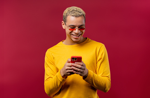 Handsome male teenager surfing internet on smartphone. Man with smile and joy on red background. Tech, success, happiness, social networks concept. High quality photo