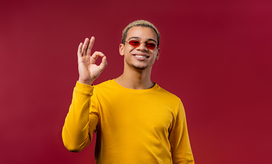 Positive man makes OK hand sign, okay gesture. Happy student guy in yellow, correct perfect choice, great deal, red studio background. . High quality photo