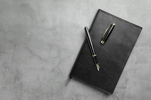 Stylish fountain pen, cap and notebook on light grey textured table, top view. Space for text