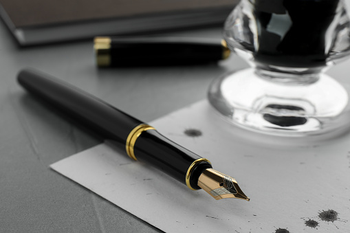 Stylish fountain pen, paper with drops of ink and inkwell on light grey textured table, closeup