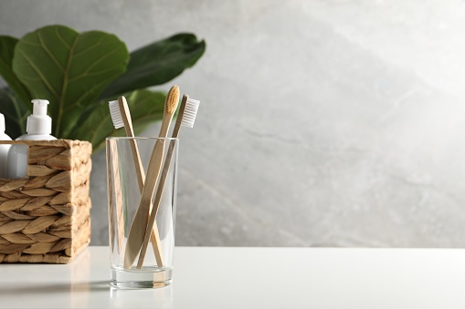 Bamboo toothbrushes in glass holder and cosmetic products on white countertop, space for text
