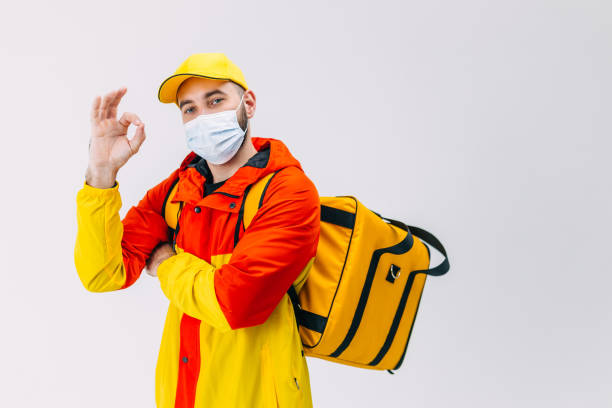 a courier in a medical mask and a yellow uniform stands with a l - manual worker thumbs up hand sign adult fotografías e imágenes de stock