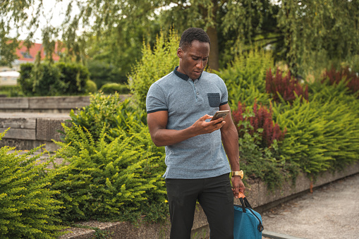 Amidst the greenery of a public park, a debonair Black male in his mid-thirties talks on the phone while confidently carrying a professional briefcase, dressed casually.