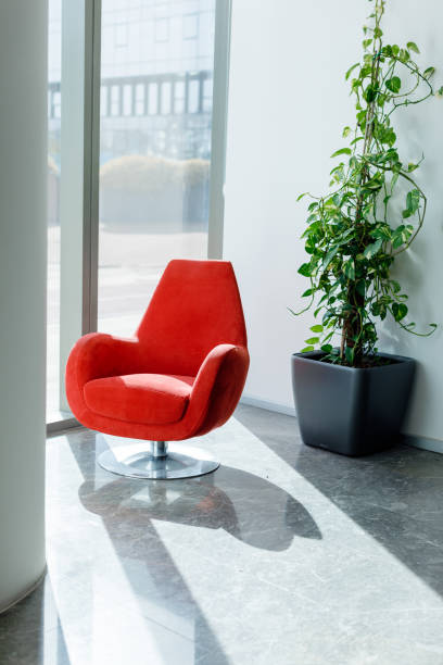 Modern Interior: Empty Red Armchair by Bright Window with Sunlight, Marble Floor. Step into a space of modern elegance with this striking stock photo, featuring an empty red modern armchair placed by a bright window. The sunlight gracefully illuminates the marble floor, creating a warm and inviting atmosphere. The scene is complemented by a green potted plant, adding a touch of nature to the contemporary interior design. This image captures the essence of sophistication and tranquility. Ideal for conveying modern interior concepts, this photo is perfect for interior design promotions, home decor publications, and social media content celebrating stylish living spaces. Modern interior, Empty red armchair, Bright window, Sunlight, Marble floor, Green potted plant, Sophistication, Tranquility, Contemporary, Interior design, Stock photo, iStock, Interior design promotions, Home decor publications, Social media content, Stylish living spaces.
