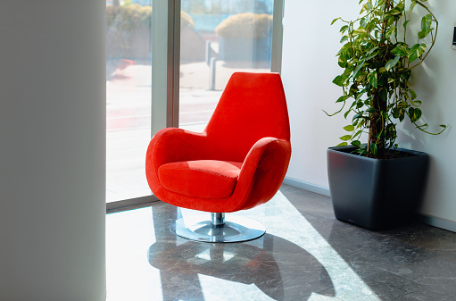 Modern Interior: Empty Red Armchair by Bright Window with Sunlight, Marble Floor. Step into a space of modern elegance with this striking stock photo, featuring an empty red modern armchair placed by a bright window. The sunlight gracefully illuminates the marble floor, creating a warm and inviting atmosphere. The scene is complemented by a green potted plant, adding a touch of nature to the contemporary interior design. This image captures the essence of sophistication and tranquility. Ideal for conveying modern interior concepts, this photo is perfect for interior design promotions, home decor publications, and social media content celebrating stylish living spaces. Modern interior, Empty red armchair, Bright window, Sunlight, Marble floor, Green potted plant, Sophistication, Tranquility, Contemporary, Interior design, Stock photo, iStock, Interior design promotions, Home decor publications, Social media content, Stylish living spaces.