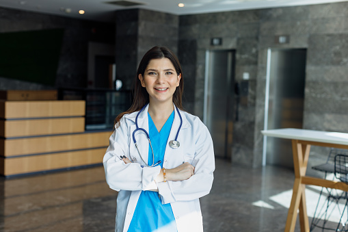 Confident Female Medical Professional in Modern Hospital Corridor. Radiate confidence and success with this empowering stock photo, showcasing a young female doctor or medical professional smiling, arms crossed, and looking at the camera in a modern hospital corridor entrance. She is adorned in a white coat and wears a stethoscope around her neck, representing her expertise and dedication to healthcare. The modern hospital setting emphasizes professionalism and commitment to patient care. This image captures the essence of a confident and successful medical professional. Ideal for conveying healthcare excellence, this photo is perfect for medical promotions, healthcare publications, and social media content celebrating dedicated professionals in the field. Confident, Successful, Young female doctor, Medical professional, Modern hospital corridor, Entrance, White coat, Stethoscope, Healthcare, Professionalism, Patient care, Stock photo, iStock, Medical promotions, Healthcare publications, Social media content, Dedicated professionals