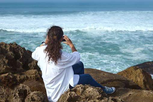 Curly haired multi ethnic pretty woman taking photo on her smart mobile phone, sitting on the rock on the Atlantic ocean beach. People. Nature. Leisure and weekend activities. View from the back