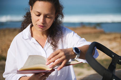 Beautiful curly Latin American young adult woman sitting near her baby stroller, leafing through the pages while reading novel on the beach. Digital detox. Literature. People. Lifestyles and hobbies