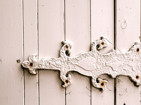 Horizontal closeup photo of a section of an old pastel pink painted wooden door with a metal heart and fleur de lys pattern hinge with rusty bolts.