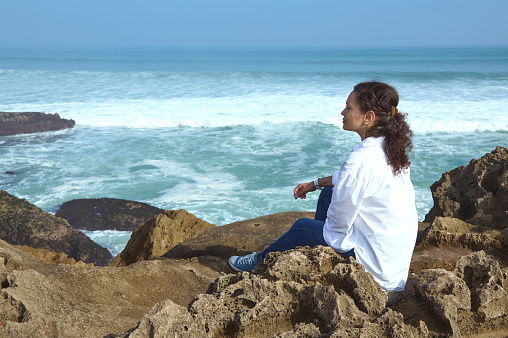 Beautiful alone woman dreamily looking into the distance, sitting on the cliff by Atlantic ocean shore and feeling connection with nature. People. Lifestyle. Leisure activity. Mindfulness