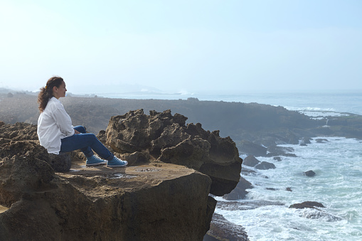 Relaxed woman in a white casual shirt and blue jeans, sitting alone on a cliff and admires the mesmerizing view of the Atlantic Ocean. People. Loneliness. Meditation. Connection with beautiful nature